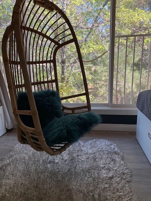 Une chaise en osier agrémentée d’un coussin vert moelleux, un tapis blanc et duveteux, et une vue sur les arbres par la fenêtre.