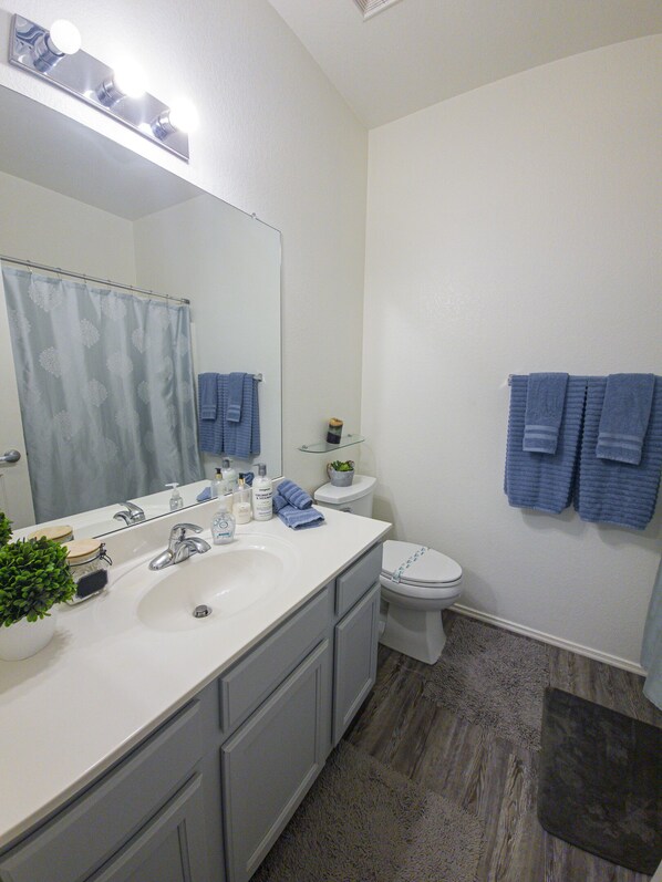 Un bagno con lavandino bianco, specchio e servizi igienici. Asciugamani blu appesi al muro e una piccola pianta sul ripiano.