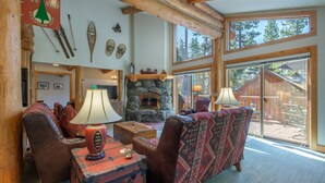 A cozy living room with a fireplace, rustic furniture, and large windows offering a view of the outdoors.