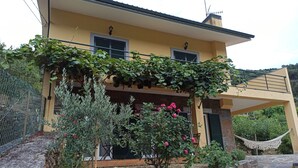 A two-story house with a yellow facade, green shutters, and a stone retaining wall.