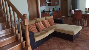 A living room with a wooden staircase, a sectional sofa with striped cushions, a kitchen area with cabinets and appliances, and a dining table with chairs.