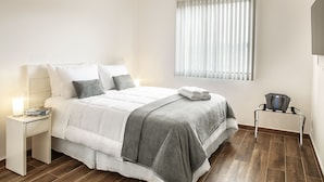 Una cama bien tendida con un edredón gris, una mesita de noche con una lámpara y una ventana con persianas.