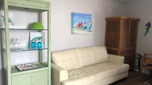 Un salon avec un canapé de couleur crème, un meuble en osier vert doté d’étagères en verre, un meuble en bois et un tableau représentant des voiliers accroché au mur.