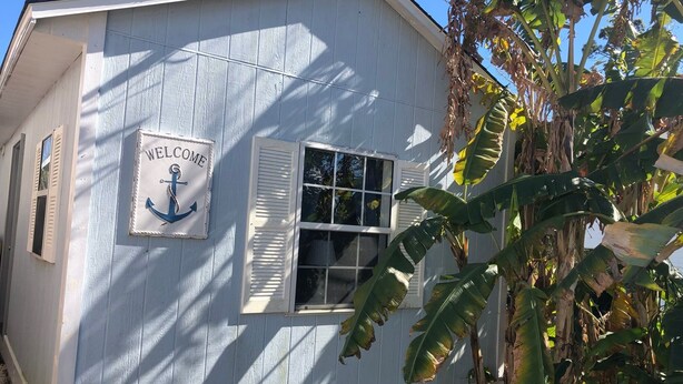 Un bâtiment blanc avec une enseigne « BIENVENUE » et un symbole d’ancre, une fenêtre avec des volets et une clôture en treillis.