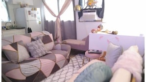 A cozy living room with a sofa, a chair, and a small table. There is a window with curtains, a radiator, and a shelf with decorative items. - Hawaiian Princess Paradise (Honolulu)