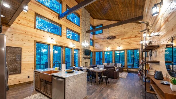 Un intérieur spacieux de chalet en bois, comprenant une cuisine, un coin repas et de grandes fenêtres offrant une vue sur la forêt.