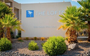 A sign for The Lofts at 1122 South 1790 West, featuring a palm tree logo and surrounded by various plants and a gravel path. - Abbys Retreat At Green Valley (St. George)