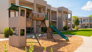 A playground with a slide and climbing wall, surrounded by a landscaped area with sand and plants. - Abbys Retreat At Green Valley (St. George)