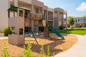 A playground with a slide and climbing wall, surrounded by a landscaped area with sand and plants. - Abbys Retreat At Green Valley (St. George)