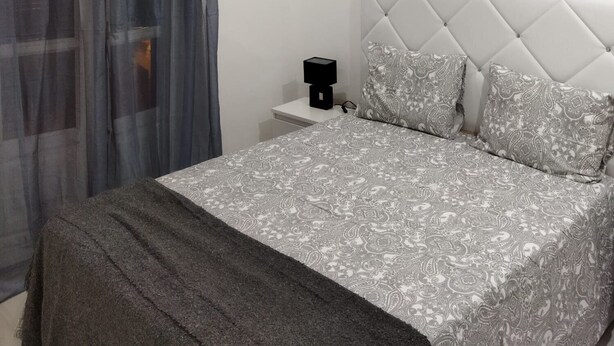 Een slaapkamer met een bed, een nachtkastje, een modern hoofdeinde en een ventilatie rooster.
