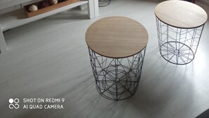 Een moderne woonkamer met een flatscreen televisie op een witte standaard, een klein plantje en twee ronde tafels met draadframe.