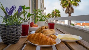 A wooden table with a croissant, a plate with pastries, a basket of flowers, and a glass of red juice on a balcony overlooking palm trees and a cityscape. - Peace & Love Flat Nazaré - Peace & Love (Nazaré)