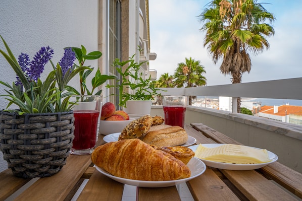 A wooden table with a croissant, a plate with pastries, a basket of flowers, and a glass of red juice on a balcony overlooking palm trees and a cityscape.