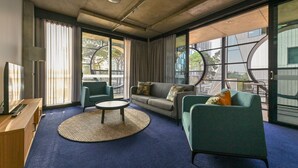 Una habitación de hotel moderna con un ventanal grande, un sofá, una mesa redonda de centro y un escritorio con lámpara.