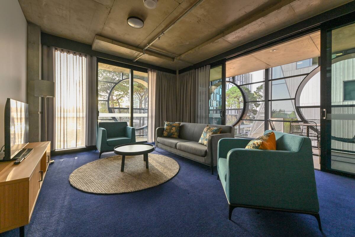 Una habitación de hotel moderna con un ventanal grande, un sofá, una mesa redonda de centro y un escritorio con lámpara.
