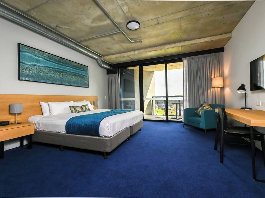 Quarto de hotel com cama, escrivaninha, cadeira, mesa de cabeceira, abajur e uma janela grande.