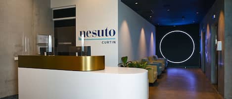 A reception desk with a modern design, a sign reading "nesuto curtin," and a hallway with seating and a circular light feature.