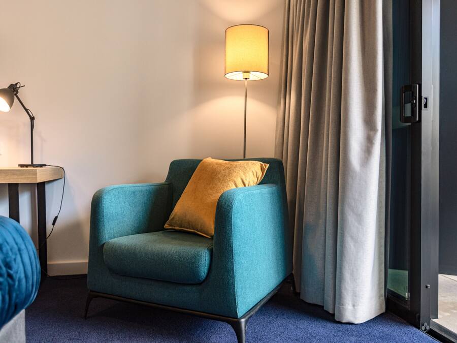 A teal armchair with a gold pillow, a floor lamp, a bed with a blue blanket, and a door with a curtain.