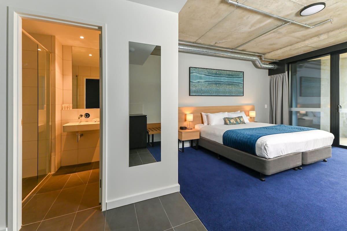Habitación de hotel moderna con una cama grande, un baño con cabina de ducha de vidrio y vistas a través de un ventanal corredera.