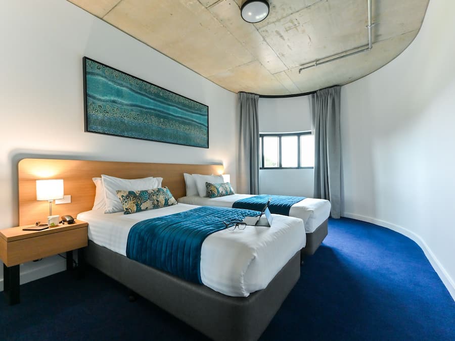 Una habitación de hotel con dos camas, una mesita de noche, una lámpara, un cuadro y una ventana con cortinas.