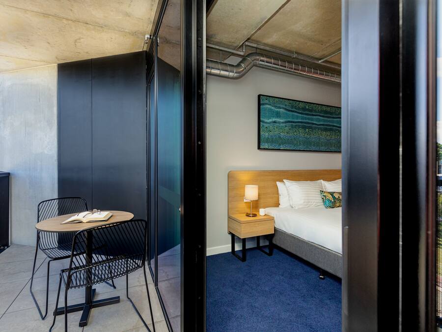 Una habitación de hotel con cama, mesita de noche, lámpara y una pequeña mesa con sillas.