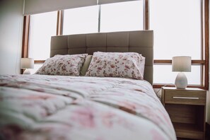 A bed with a floral bedspread and matching pillows, a wooden nightstand with a lamp, and a window with blinds.
