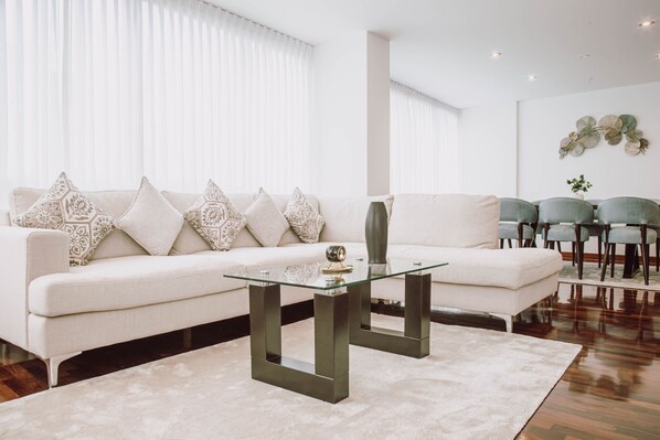 A modern living room with a white sofa, patterned cushions, a glass coffee table, and a green vase.
