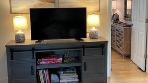 Un salon avec un téléviseur à écran plat posé sur un meuble en bois, flanqué de deux lampes décoratives. Sous le téléviseur, on trouve une bibliothèque remplie de divers livres et un vélo.