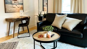 Un salon avec un canapé en cuir noir, une table basse en bois, un bureau avec une chaise, une plante en pot et un tableau représentant une scène de plage.