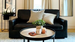 Un salon moderne avec un canapé en cuir noir, deux coussins, une table basse ronde agrémentée d’objets décoratifs, une plante en pot et un tableau encadré accroché au mur.