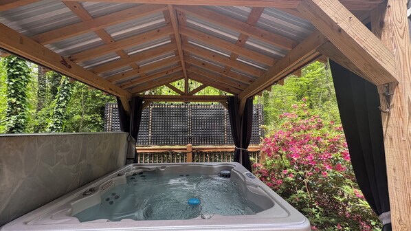 A hot tub under a wooden pergola with a view of greenery and flowers.