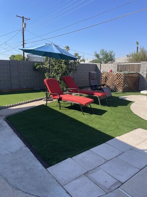 A backyard with artificial grass, two red lounge chairs, a blue umbrella, and a wooden fence.