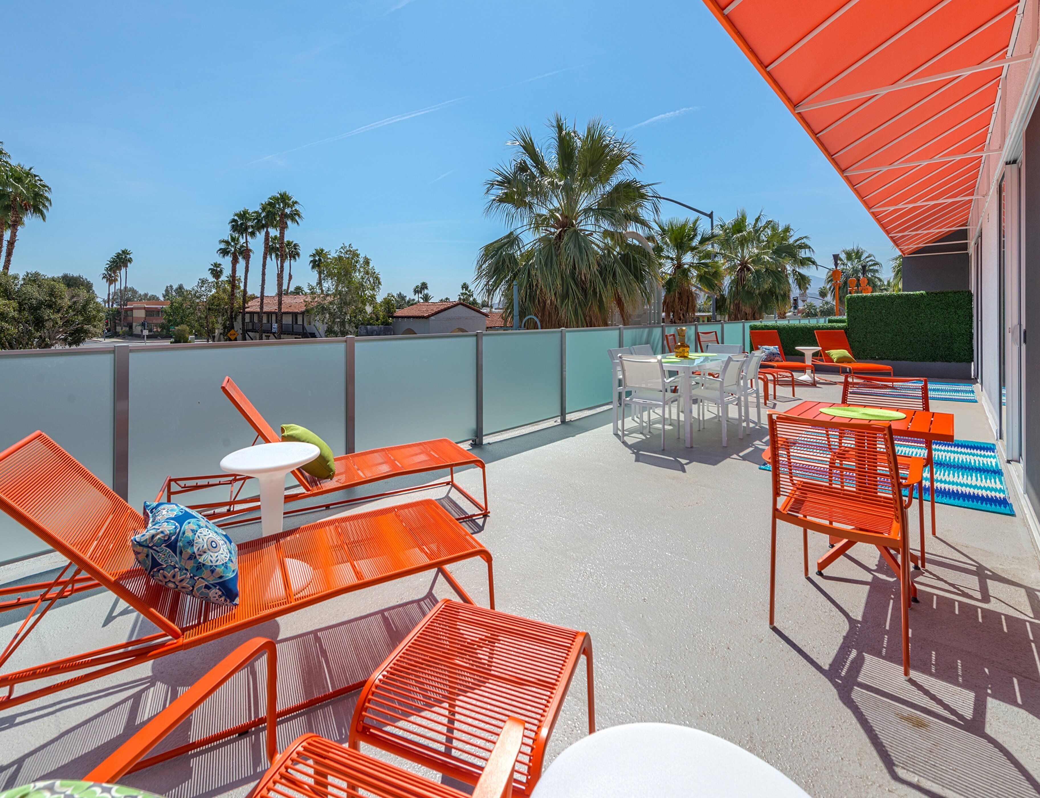 A rooftop terrace with orange metal chairs and tables, a small round table, and a view of palm trees and buildings in the background.