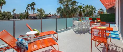 A rooftop terrace with orange metal chairs and tables, a small round table, and a view of palm trees and buildings in the background.