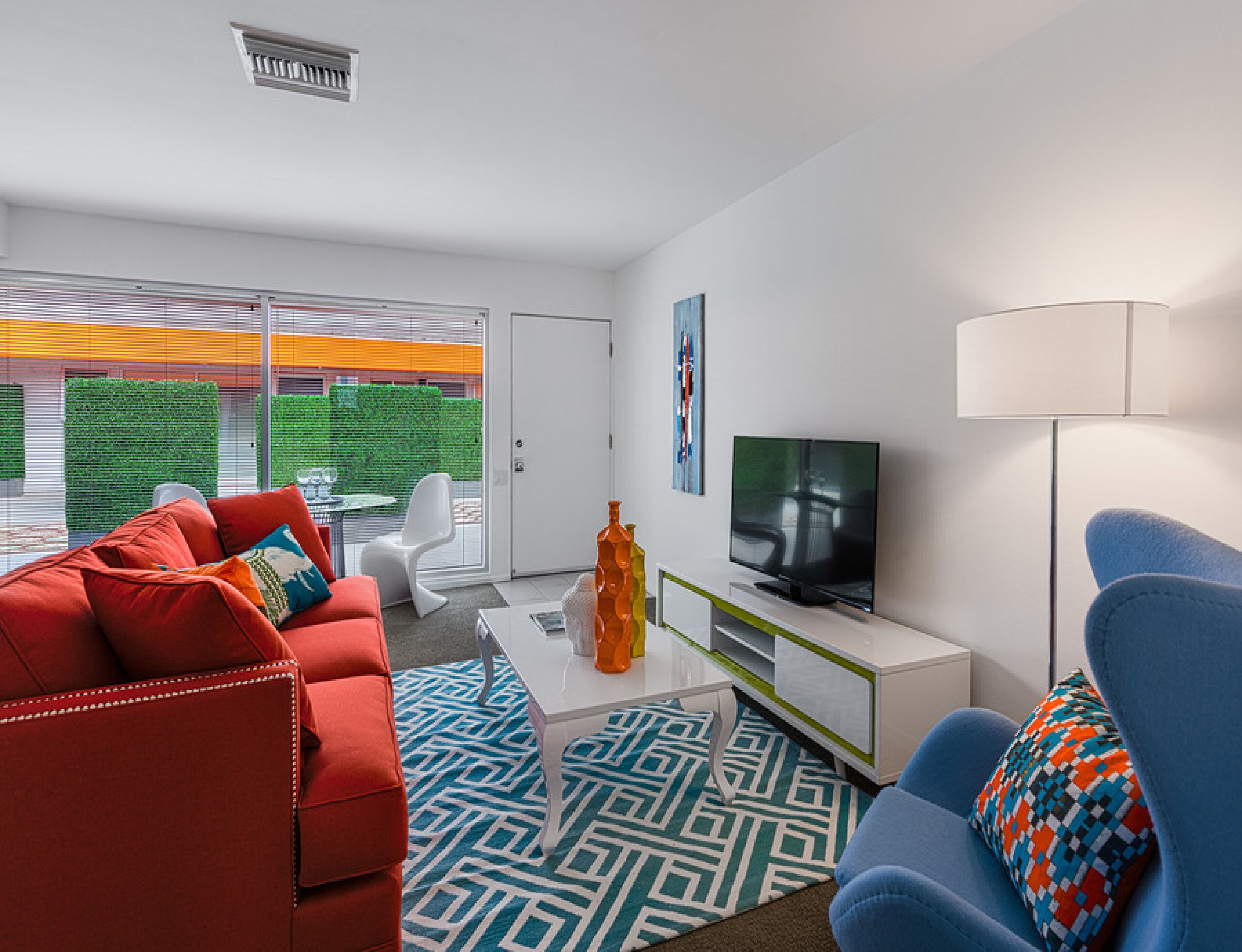 A modern living room with a red sofa, blue armchair, and a white coffee table. A television is mounted on the wall, and there is a window with blinds.