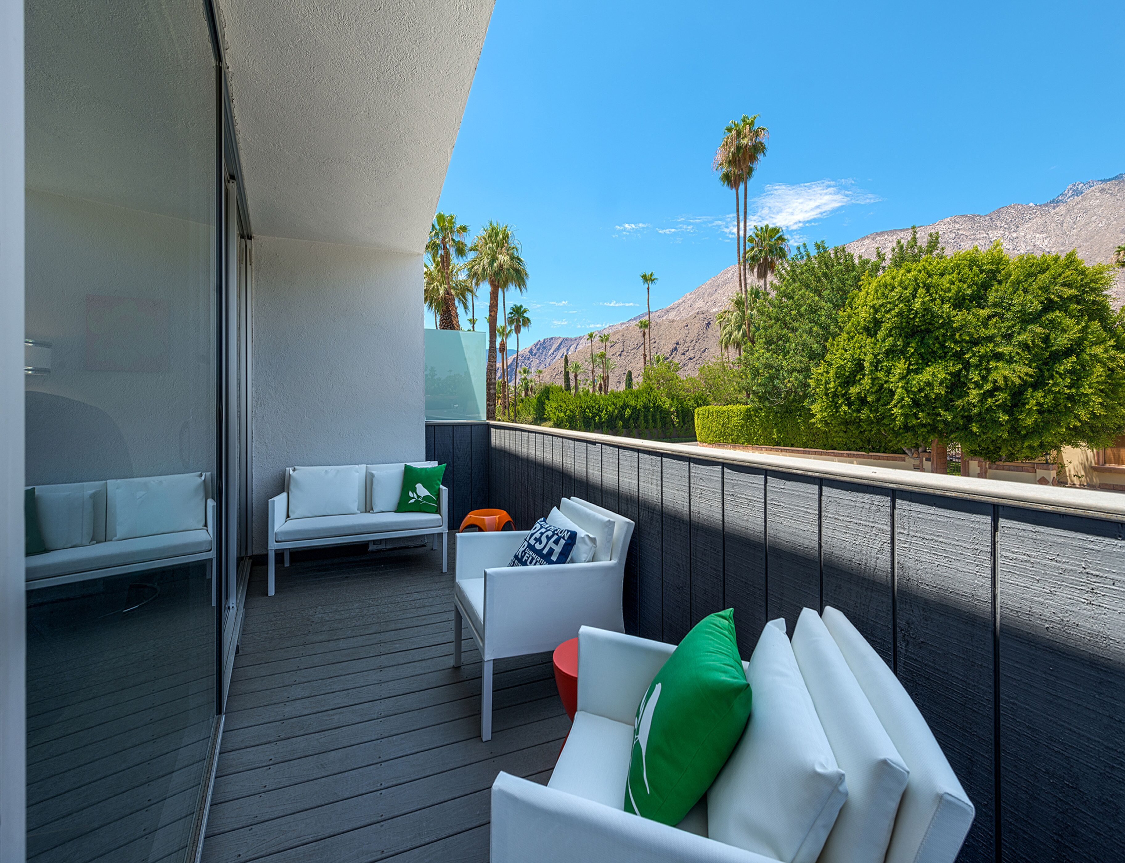 A balcony with a white sofa and two chairs, a small orange stool, and a green pillow. The balcony overlooks a mountain and palm trees.