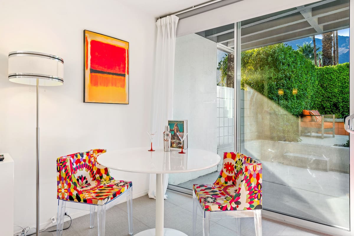 A modern room with a round white table, two colorful chairs, a floor lamp, and a large window offering a view of greenery and mountains.