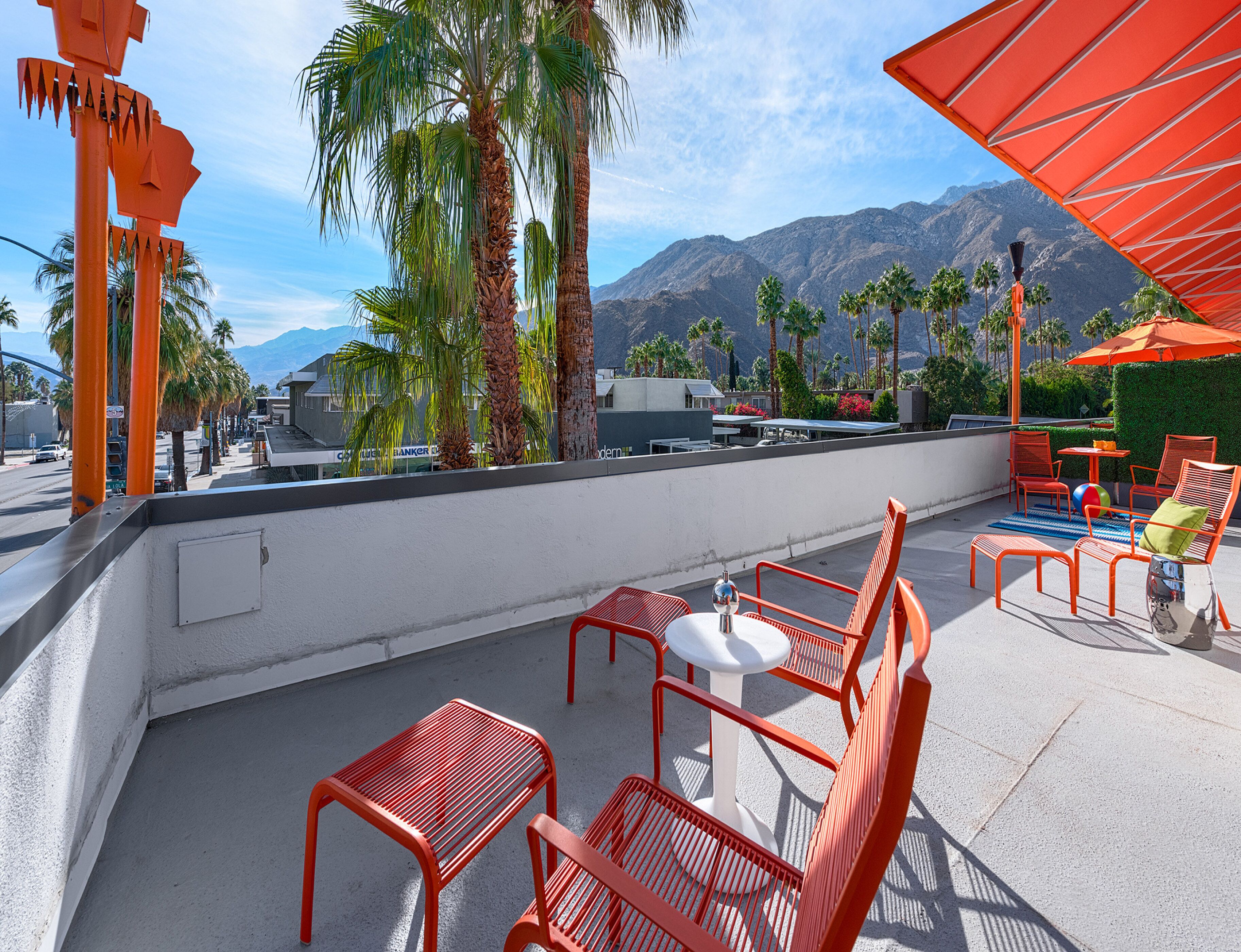 A rooftop terrace with red metal chairs and tables, palm trees, and a mountain view.