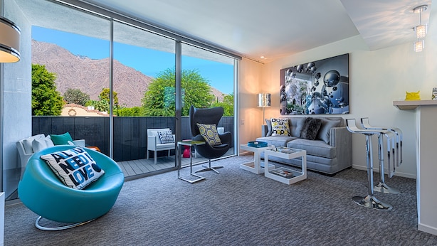 A modern living room with a large sliding glass door leading to a balcony, a teal bean bag chair, a grey sofa, and a coffee table.