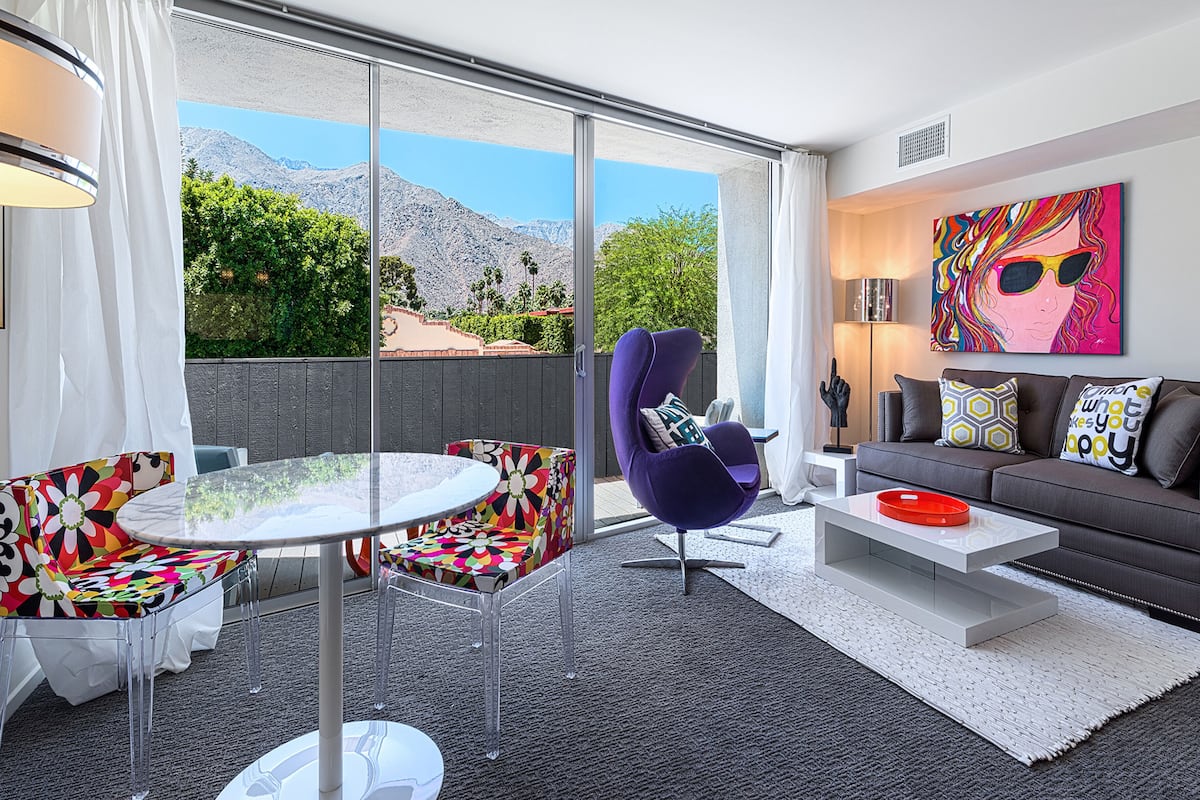 A modern living room with a large sliding glass door leading to a balcony, a round table, a purple armchair, a sofa with patterned cushions, and a colorful abstract painting.