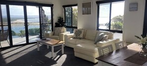 A spacious living room with a white sofa, a coffee table, and a view of a body of water through large windows. - Sunset Beach House 1770 Level 3 (Seventeen Seventy)