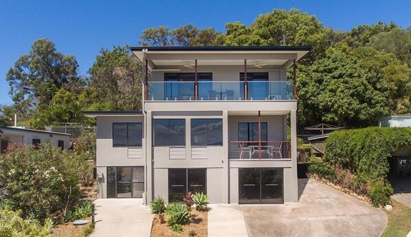 Uma casa moderna de dois andares com uma varanda no andar superior, rodeada por vegetação.