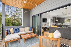 Un salon moderne avec un canapé, une table basse et des fauteuils, une cuisine à l’arrière-plan et une grande fenêtre donnant sur des arbres.