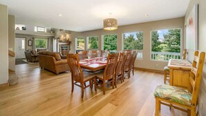 A spacious dining area with a wooden table and chairs, large windows offering a view of greenery, and a cozy living area with a sofa and fireplace.