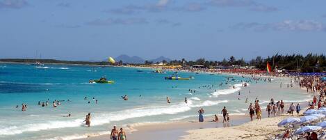A crowded beach with people walking and swimming, and a parasail in the sky.