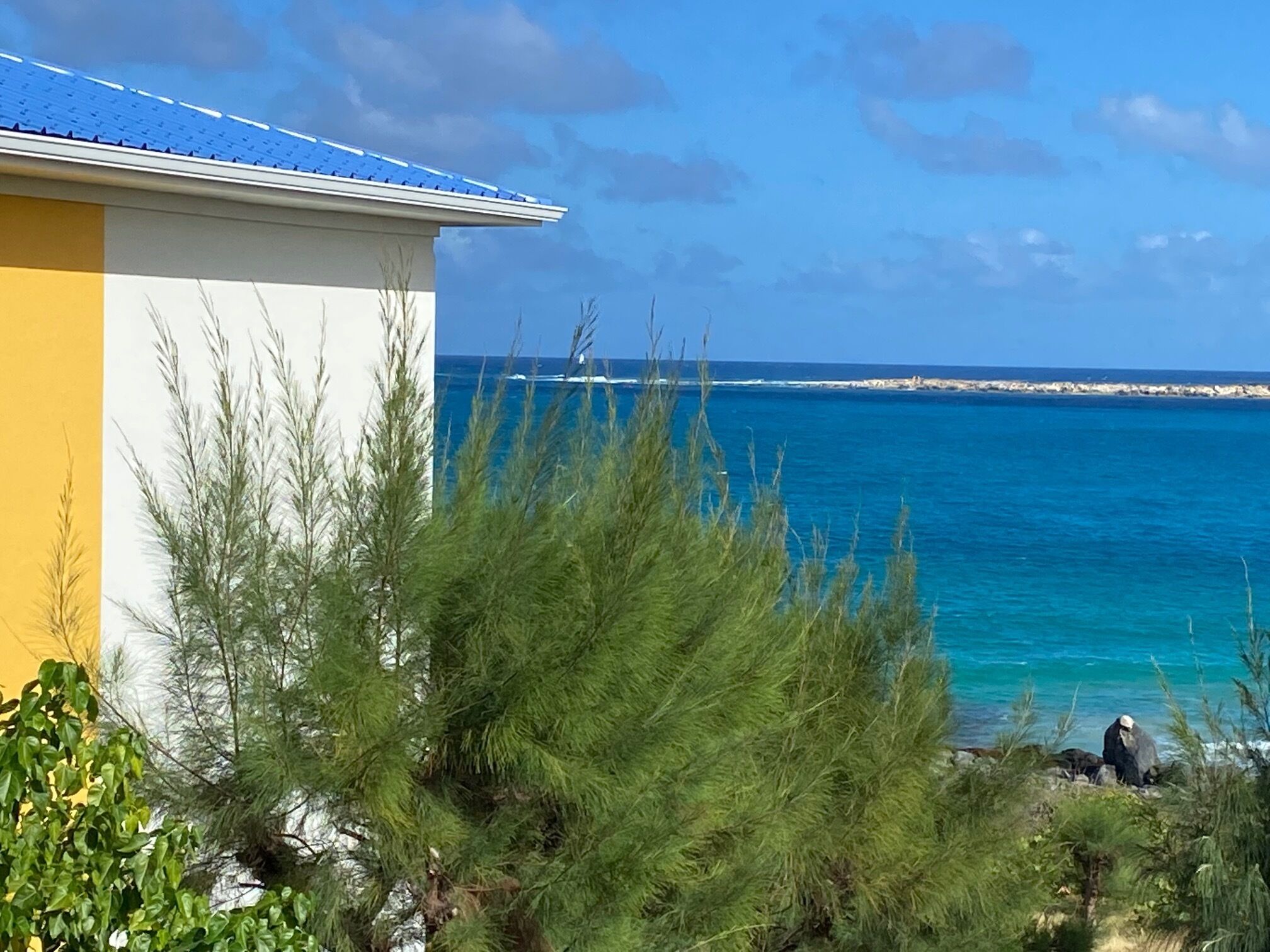 A coastal view with a building featuring a blue roof and white walls, overlooking turquoise waters and a rocky shoreline.