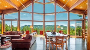 Un'ampia stanza con soffitto alto, grandi finestre con vista sulle montagne, zona pranzo con tavolo e sedie, e salotto con divani e poltrone.