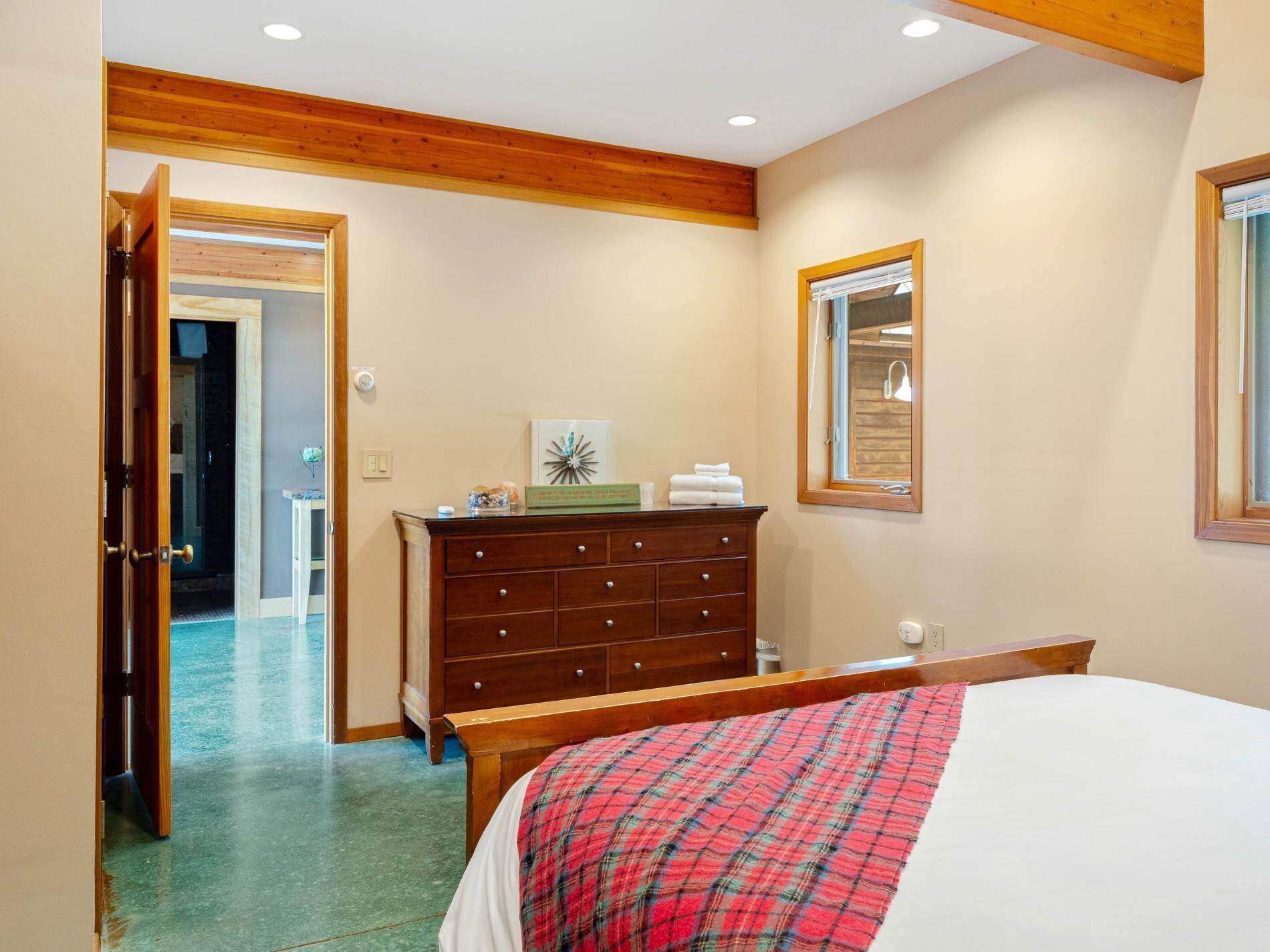 Un dormitorio con un aparador de madera, una cama con manta escocesa, un espejo y una puerta que da a otra habitación.