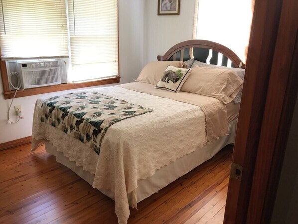 A bedroom with a bed, a window with blinds, an air conditioning unit, and a wooden floor.
