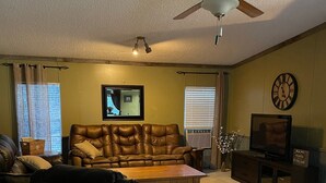 A living room with a brown leather sofa, a black leather chair, a wooden coffee table, a ceiling fan, a television, and a clock on the wall. - Lake Home  (Pontiac)